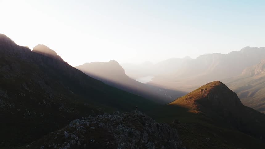 This drone shot captures a majestic rugged mountain valley bathed in the bright light of either sunrise or sunset sunlight streaks through the hazy air illuminating the peak of a central conical hill while the surrounding dramatic slopes remain in dee