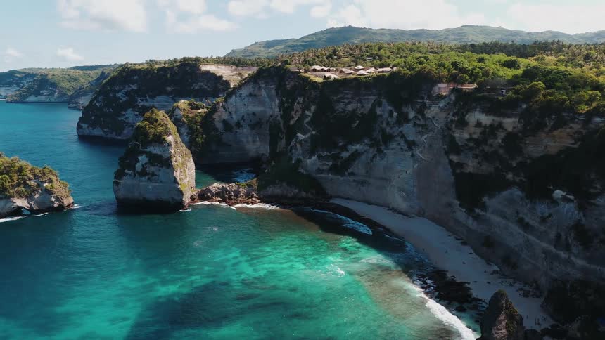 An aerial view of a small coastal beach next to a large cliff and the water is crystal clear