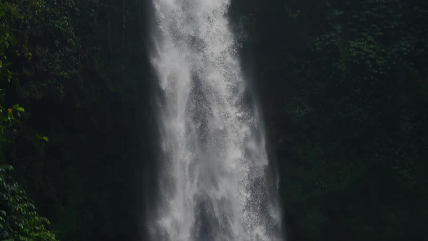 A tall powerful waterfall cascades down a steep cliff face surrounded by dense dark green tropical foliagethe camera slowly pans down to reveal the misty base of the falls where the water crashes into a pool with a person briefly visible near the r