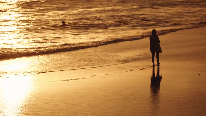 Person walking on wet sand with elongated shadow at sunset facing the sea