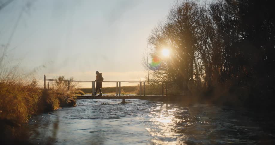 A person crossing a small wooden bridge over a fastflowing river on a sunny morning