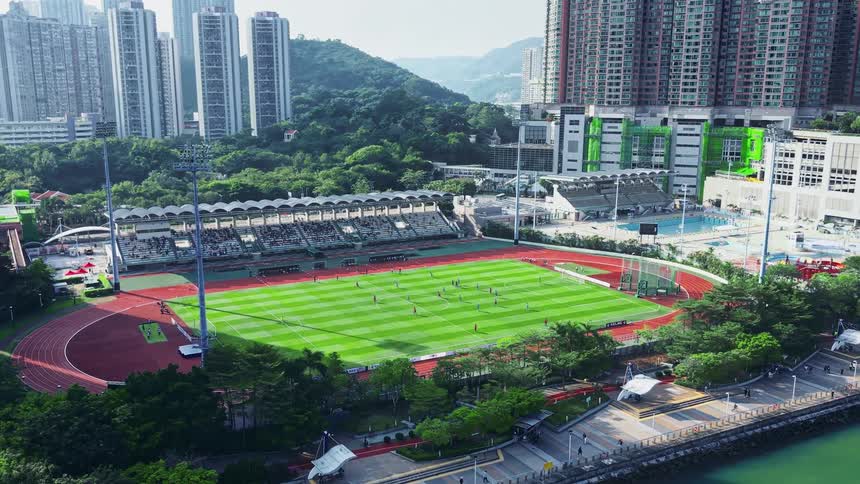 A football field in the city hosts a sporting event at midday with fans watching and cheering from the stands