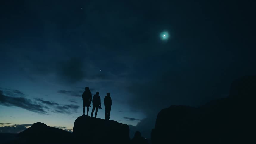 Three figures are standing close together on a rocky elevation looking up at a bright crescent moon and possibly a star or planet in the dark skythe scene is dimly lit emphasizing the silhouettes of the people and the stark outline of the landscape a