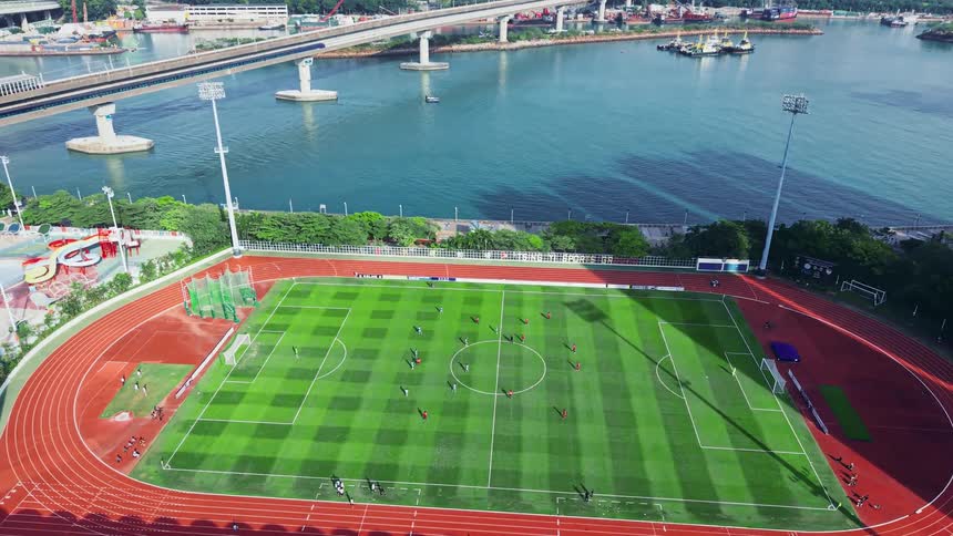 An aerial view shows a soccer stadium nestled near a wide body of water with a bridge and dense highrise buildings climbing the surrounding hills