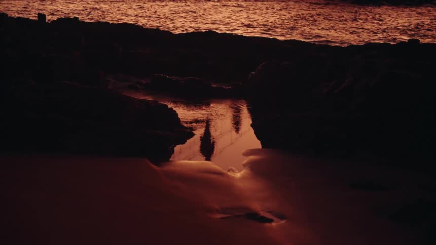 A low angle shot shows the dark silhouette of a person reflected in the water of a small pool among the rocks at sunset with the orange ocean visible in the background