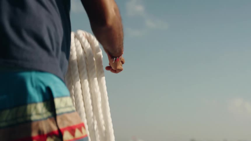 A person shown from the chest down is coiling or handling a thick white nautical rope on a boatthe bright sky and water suggest a sunny day at sea and the person is wearing a dark tshirt and patterned swim trunks