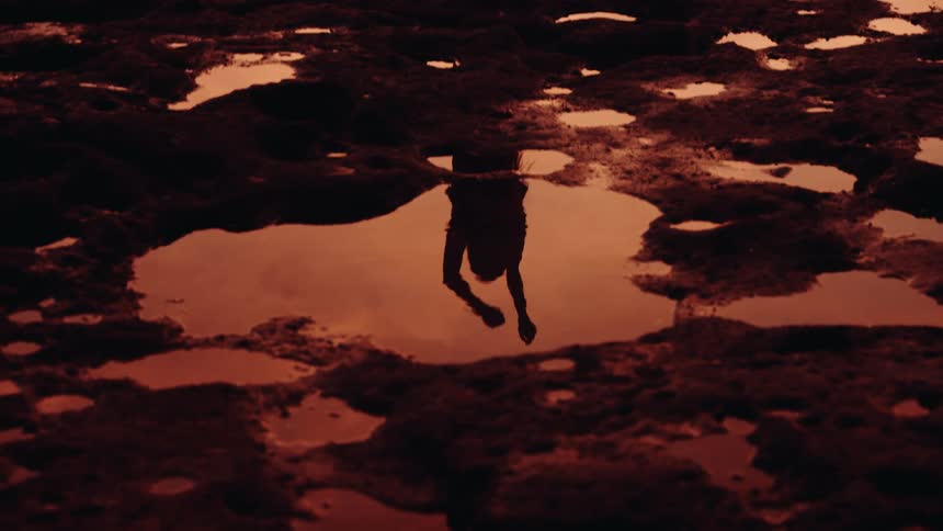 An overhead shot focuses on a dark uneven ground covered in small puddles that reflect an orange sky as the dark silhouette of a person walks directly over the center puddle