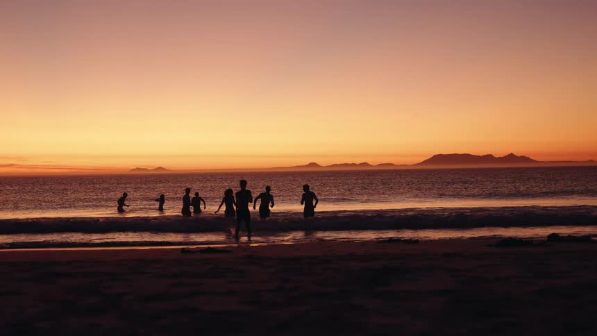 The silhouettes of several people standing and playing in the shallow ocean surf near the dark beach with the entire scene backdropped by a vibrant orange and yellow sunset over the water and distant mountains