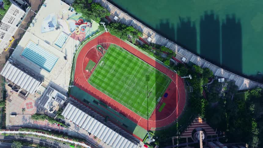 An aerial view descends slowly zooming in on a brightly lit sports complex nestled between highrise buildings and a body of water the complex features a checkered green turf soccer field surrounded by a red running track with a swimming pool and other