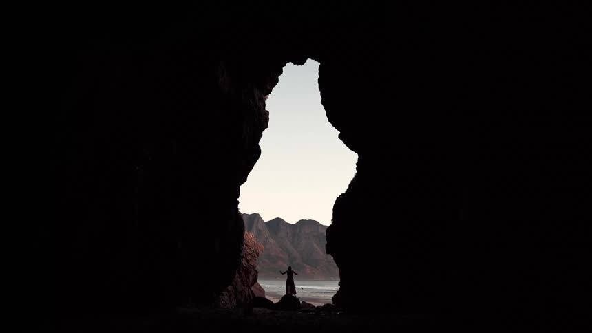 View from a hole in a cave of a woman walking on rocks on the beach as waves roll in to shore