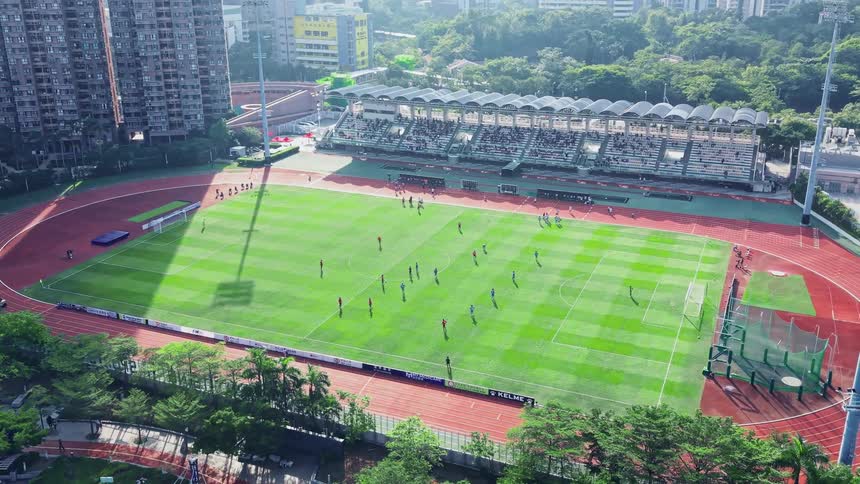 An overhead drone shot slowly pulls out to reveal an athletic stadium complex featuring a striped green football pitch and a surrounding red running track nestled within a densely populated urban area a small crowd is visible in the main covered grands