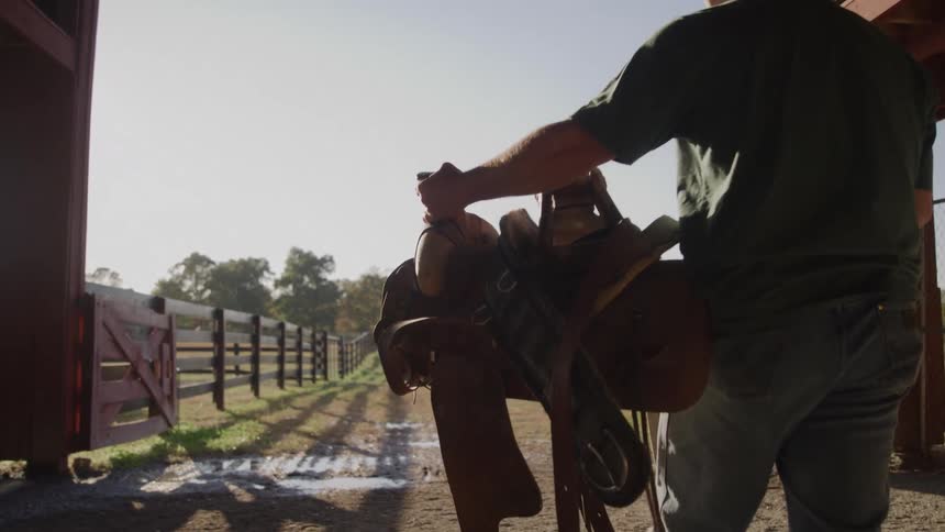 A person seen from behind is standing inside a barn or stable doorway holding a saddle the background shows a sunlit dirt path lined by wooden fences indicating a rural or ranch setting