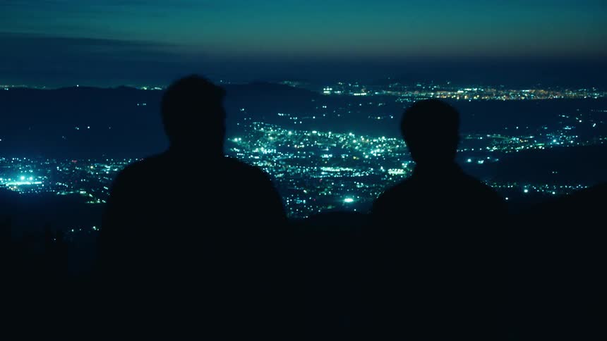 Silhouette of two men talking on top of a hill with city lights in the background outdoors during night time
