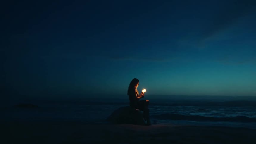 A wide shot captures the dark silhouette of a woman sitting on the shoreline against the deep blue night sky and ocean holding a small brightly glowing orange light source in her hands near her face