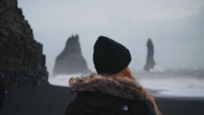 The video shows a person from behind wearing a black hat and a furtrimmed hood looking out at a dramatic black sand beach in the background the sea crashes against the shore and distinctive basalt sea stacks rise out of the misty water