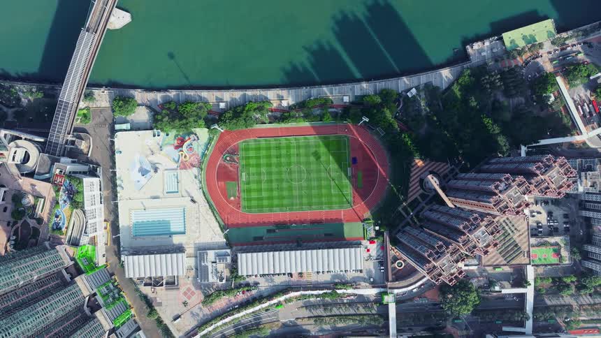 An aerial view of a riverside sports complex featuring a track and soccer field is surrounded by high rise residential buildings
