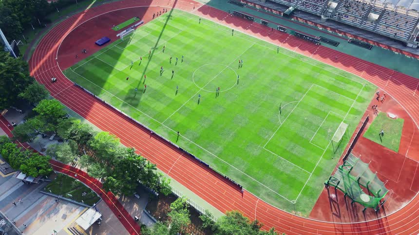 An aerial shot slowly zooms in and tilts down onto a sports stadium which features a striped green football field surrounded by a red running track the stadium is nestled between a waterfront promenade and dense modern highrise apartment buildings