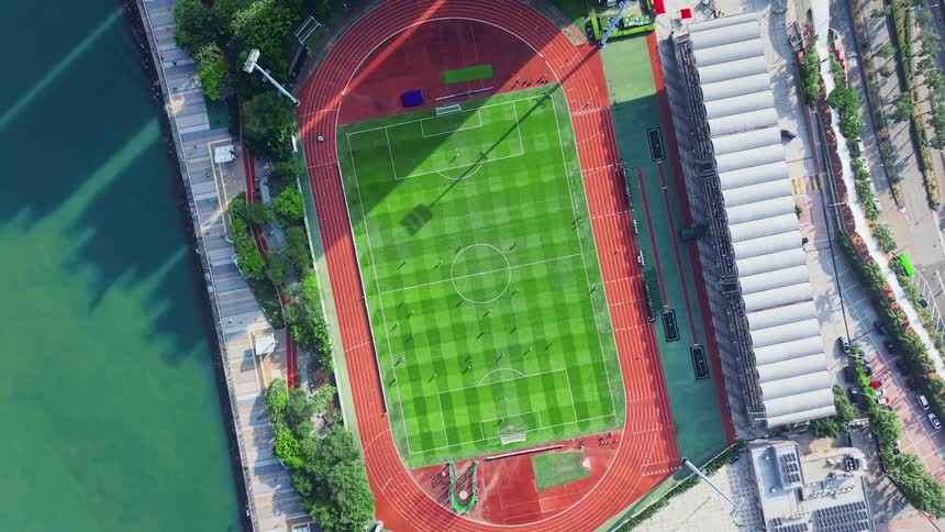 This is an extreme overhead aerial shot of a bright green football or soccer stadium surrounded by a vibrant reddishbrown running trackthe stadium is situated right next to a body of green water and there are small figures of people playing on the f