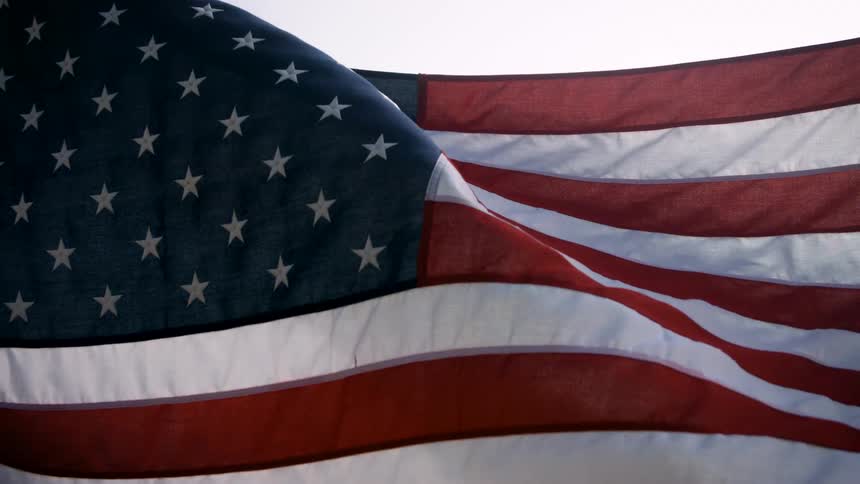 A large american flag waves slowly in the breeze its red and white stripes rippling gently alongside the field of white stars against a bright sky