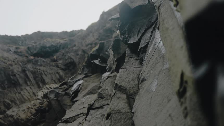 The video shows a closeup dynamic shot looking up the massive wet and rugged face of a dark rock cliff or quarrythe texture of the rock is prominent with steep angles and many fractured irregular surfaces leading up toward a foggy or overcast sk