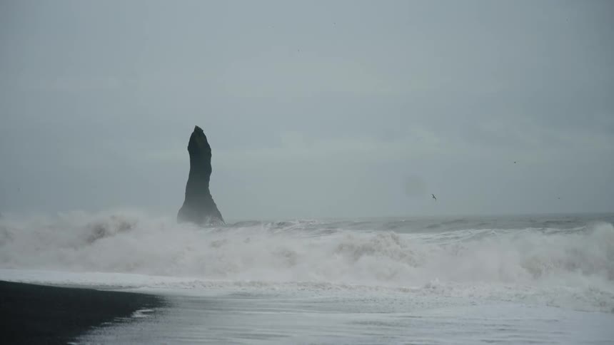 The video features a dramatic moody scene of a tall dark sea stack rising sharply out of the ocean against a grey and overcast sky powerful white waves crash repeatedly around the base of the rock formation and roll onto a black sand beach in the fore