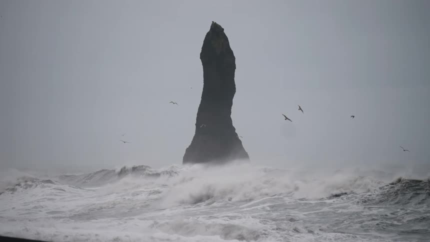 A towering sea stack rises amid crashing waves under a stormy sky with birds flying overhead