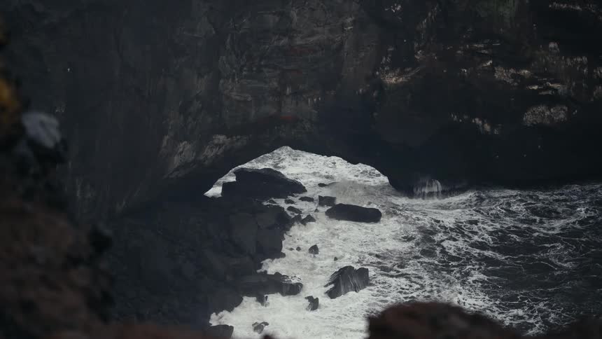 A static highangle shot looks down into a dark rocky cove or sea cave opening at the base of a high cliff where strong ocean waves crash against the rock formations creating white foam and turbulent water within the confined natural archway