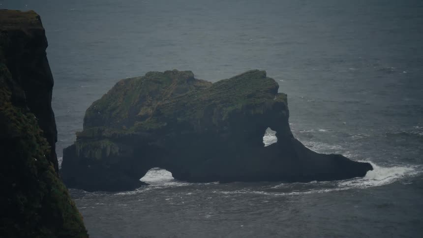 Birds are flying around a rock formation just off a coast with mountains