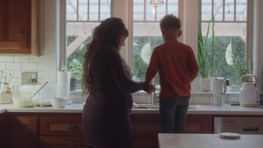 This video shows a pregnant woman with long dark wavy hair and a young boy with curly hair standing at a kitchen sink they are facing away from the camera looking out a large window as they appear to be washing or preparing something together