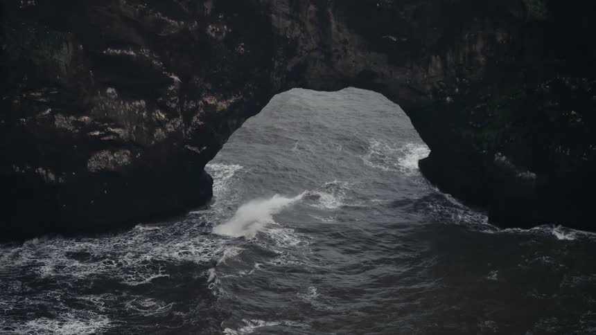 An aerial view shows powerful frothy ocean waves crashing into the dark opening of a large natural sea cave carved into a steep cliff face