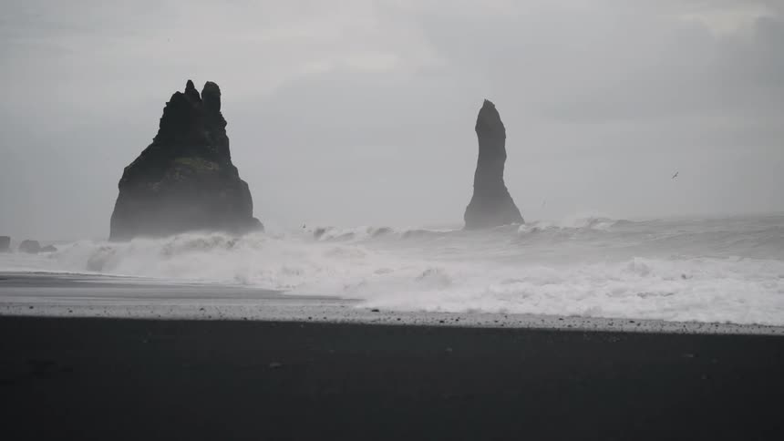 A large waves crash on a black sand beach with two sea stacks under a moody sky