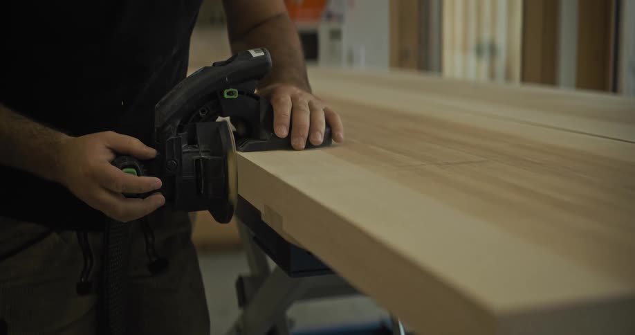 A person uses a circular saw to cut a thick wooden plank in an indoor workshop