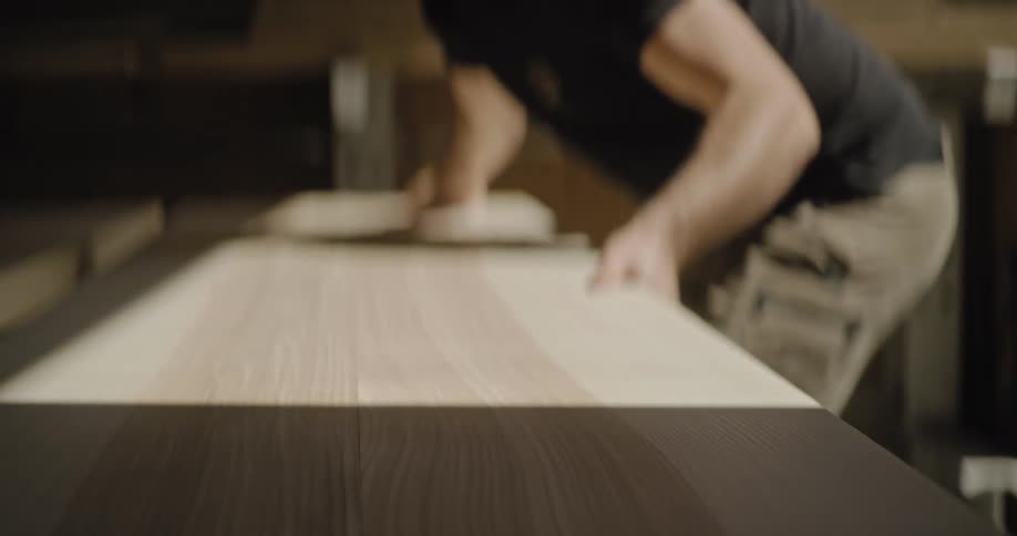 A woodworker wearing a dark tshirt and light trousers slides a long piece of lightcolored finished wood across a surface aligning it with other planks the action takes place in a dimly lit workshop focusing on the woods smooth grain