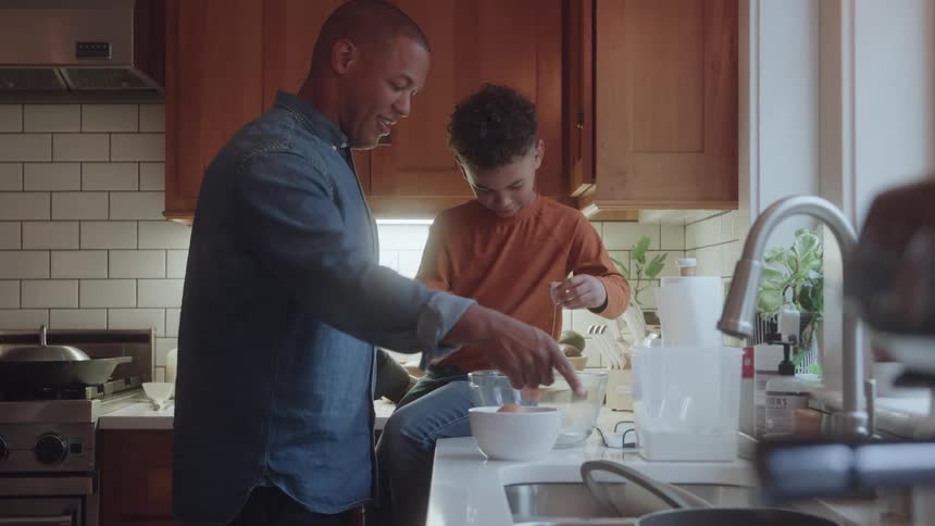 A boy is cracking eggs into a bowl as his father watches and cleans up