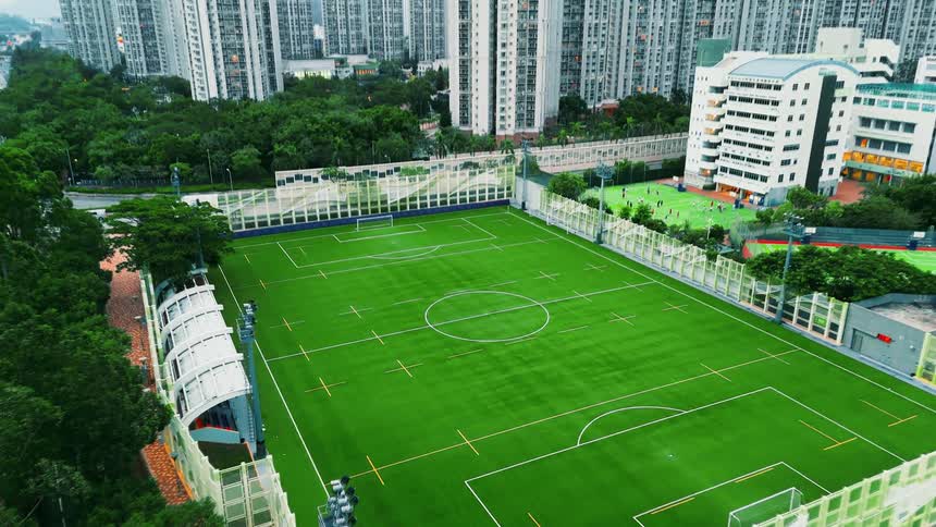 An overhead view captures a bright green synthetic turf football pitch surrounded by highdensity residential towers and a multistory school building the urban environment is dominated by the tall densely packed apartment blocks looming over the recre