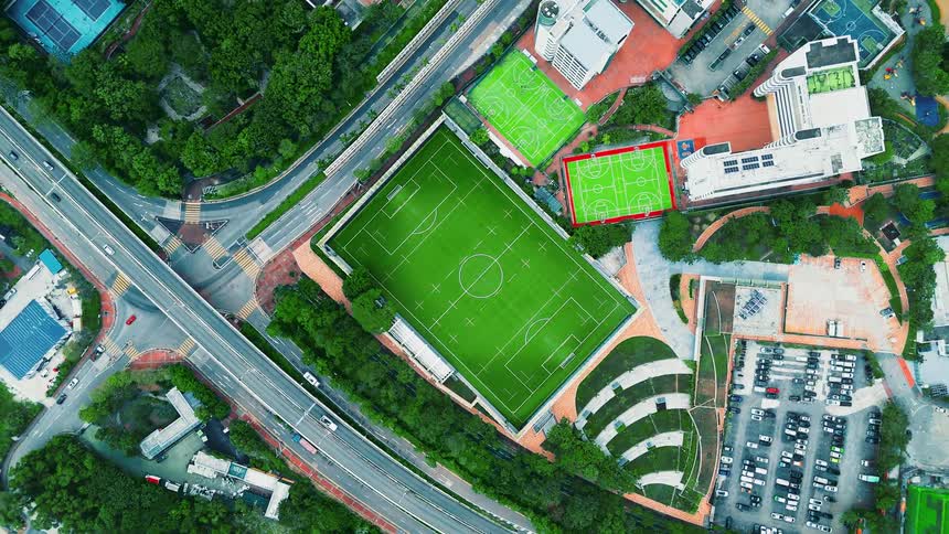 This is an aerial drone shot of a multisport complex surrounded by urban infrastructure featuring a large bright green soccer field and two smaller courts above it the complex is bordered by dense green trees a large parking lot and busy roadways