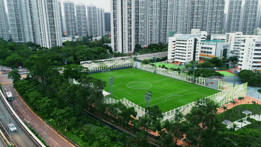 An aerial shot captures a modern caged artificial turf football pitch bordered by a busy highway and lush green trees the pitch is dramatically contrasted against a dense background of numerous tall highrise residential towers and a lower school buil