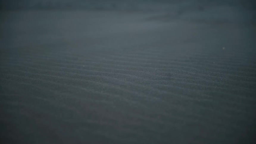 This video shows a dark textured surface likely sand or fine dirt covered with subtle ripples created by wind or water the colors are muted and monochromatic giving the scene a somber or cold atmosphere