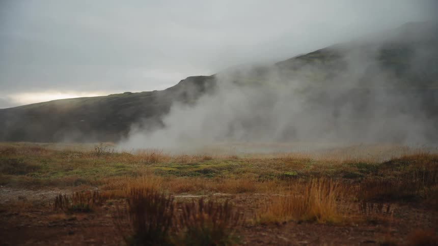 This clip shows a wide shot of a misty geothermally active landscape likely in a volcanic region like icelandthick plumes of steam rise vigorously from the ground obscuring the base of a dark grassy hillside under a moody sky with dry brown and y