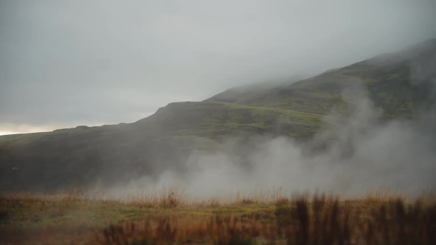 Mist drifts from a grassy field obscuring the base of a green hillside under an overcast sky