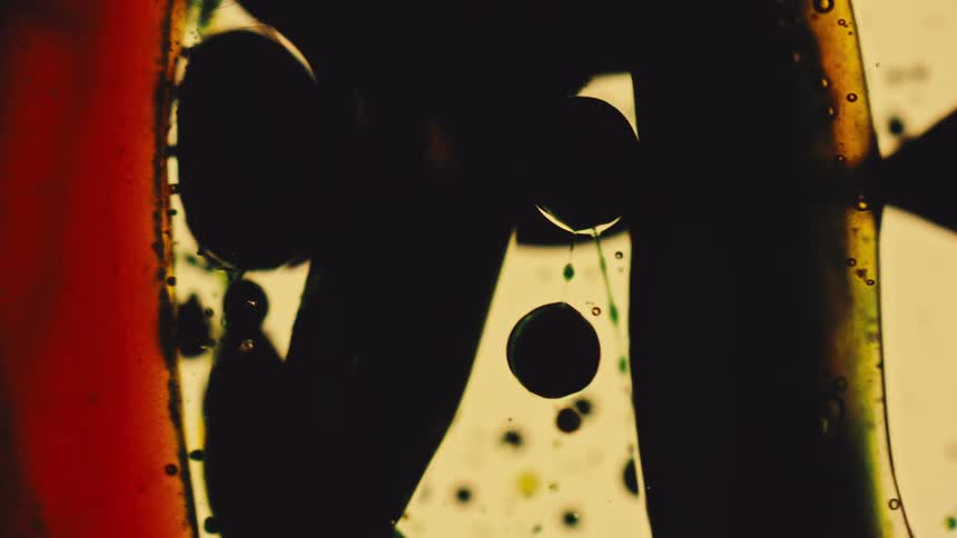 The video shows an abstract macro shot of dark fluid shapes like oil or ink blobs moving and merging against a lighter yellowish backgroundto the left a vertical stripe of reddishorange color frames the scene adding a strong color contrast to
