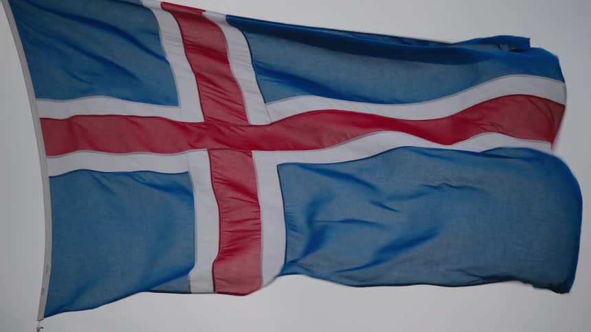 An icelandic flag flies on a pole at midday billowing in the wind against the open sky