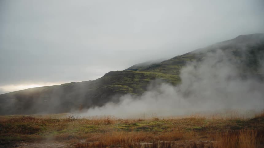 This is a wide shot of a geothermal landscape with steam vigorously rising from the ground in the foregroundthe steam originates from a dry goldenbrown grassy field set against a dark green mistshrouded mountain slope under an overcast sky