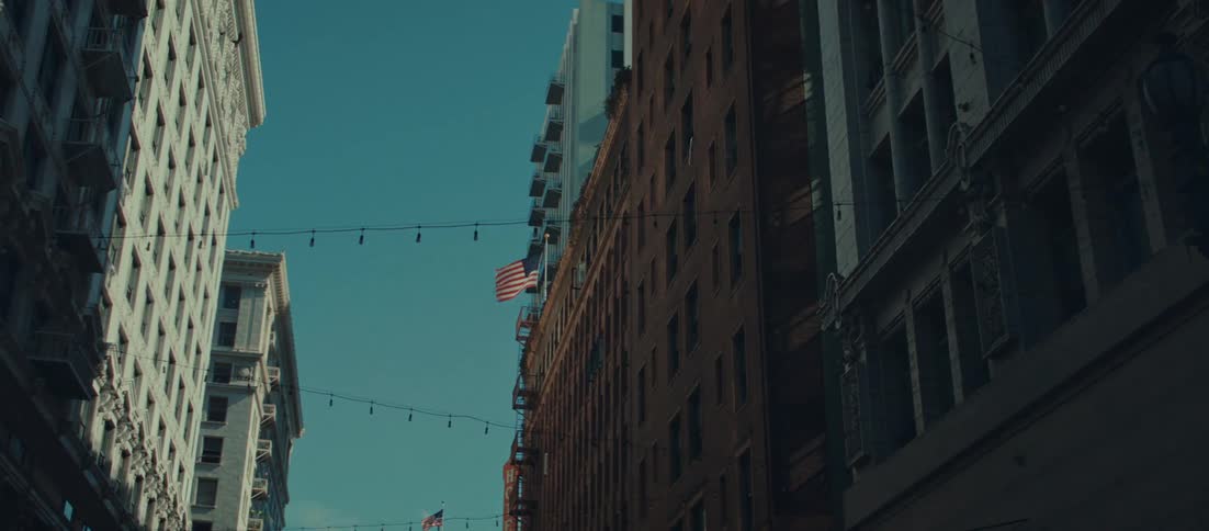 The facades of multiple tall buildings lining a street in an urban setting with string lights strung across the space between the structures and american flags attached to the darker bricksided building under a clear blue sky