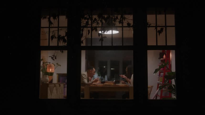 Cozy nighttime scene with two people dining in warmly lit room viewed through large window