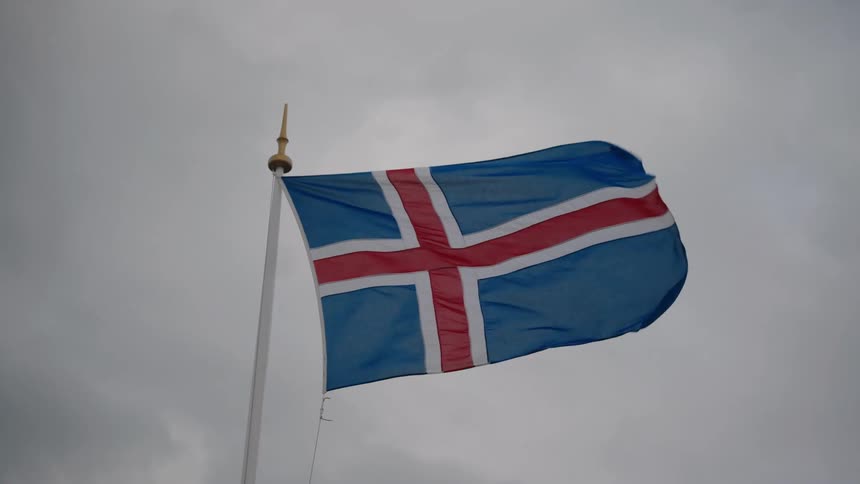 This video clip features the flag of iceland which is blue with a red cross bordered in white waving on a flagpole the background is an overcast gray sky suggesting a cloudy day