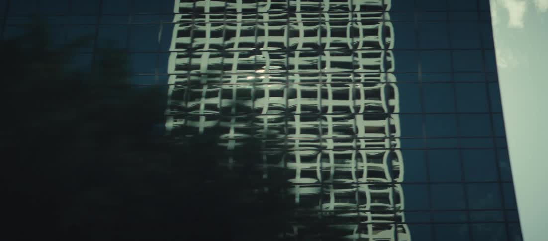 A moving shot shows a distorted building reflection on a glass skyscraper followed by a bright lens flare and a view of the blue glass facade with trees
