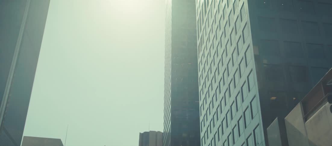 A vertical pan shot travels down the exterior of a dark modern skyscraper in a city setting with the camera pointing almost straight up at the start the bright washedout sky with strong lens flare dominates the upper part of the frame before the came