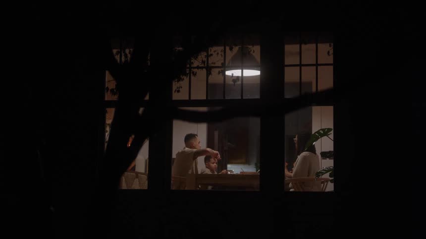 Viewed through a dark window from outside a family sits together at a dining table sharing a meal in the warm light of a hanging lamp