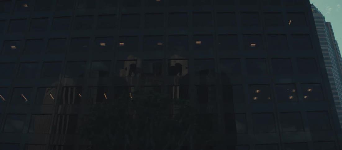 The video starts with a closeup of a dark modern corporate building facade featuring a grid of reflective glass windows the camera pans slowly to the right and zooms out revealing other tall buildings in the background under a bright sky characteris